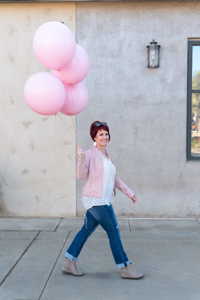 Brand photo taken by Robin Collette Photography. Prop idea for personal branding photoshoot Female wearing pink jacket and walking down a sidewalk holding 4 large pink helium balloons by attached strings.
