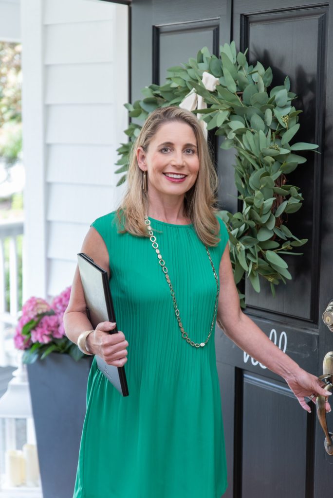 female realtor in green dress standing in a home doorway holding a clipboard